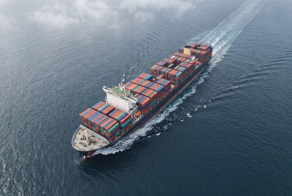 Luftaufnahme eines großen Containerschiffs auf dem Meer, beladen mit bunter Fracht, das eine weiße Heckwelle erzeugt.
