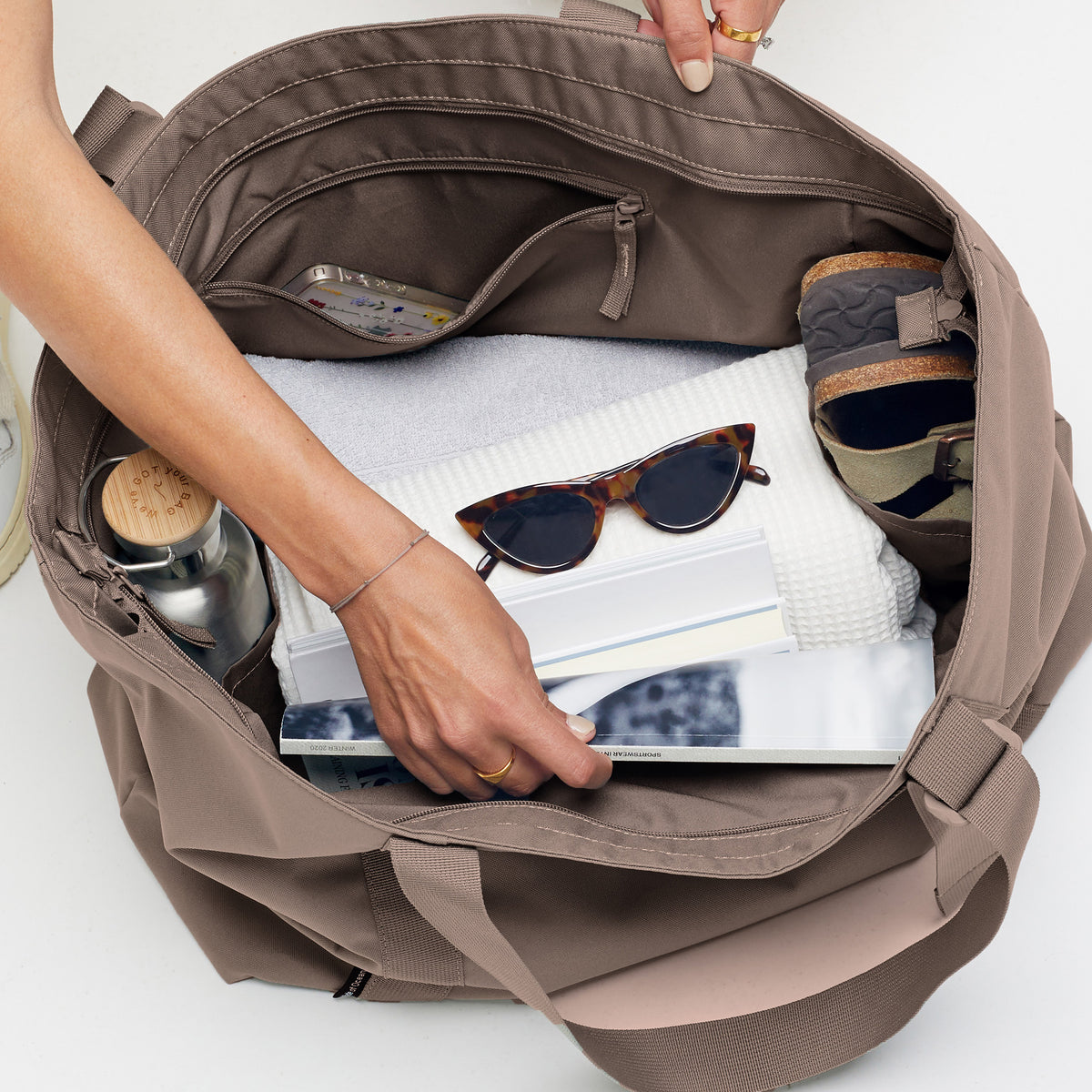 TOTE BAG Tasche in seal (dunkler Beigeton) wird von einer Frau mit Gegenständen befüllt.