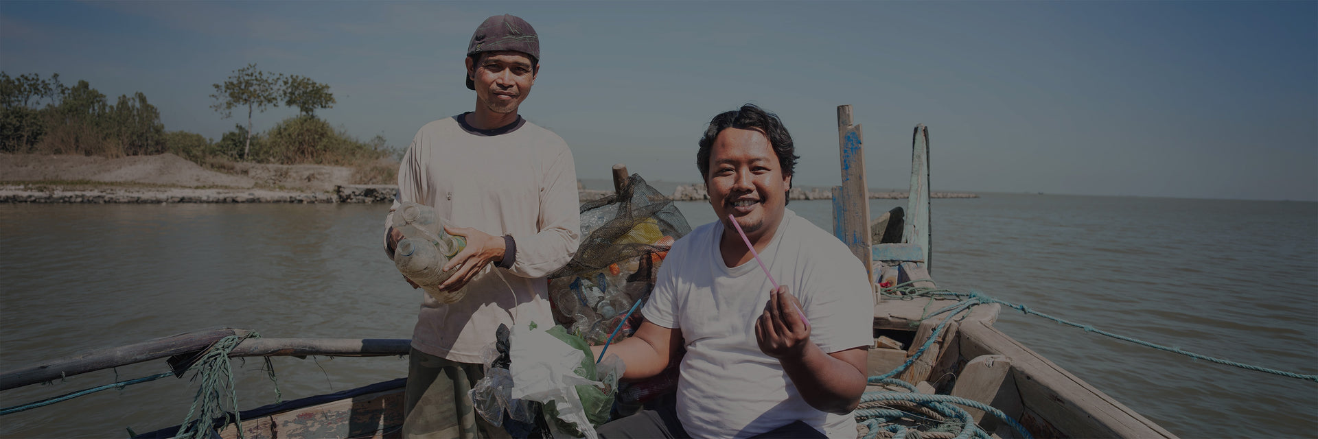 COMMUNITY LEADERS Accessoires: Zwei Männer sammeln Plastikmüll am Wasser, einer hält einen pinken Strohhalm.