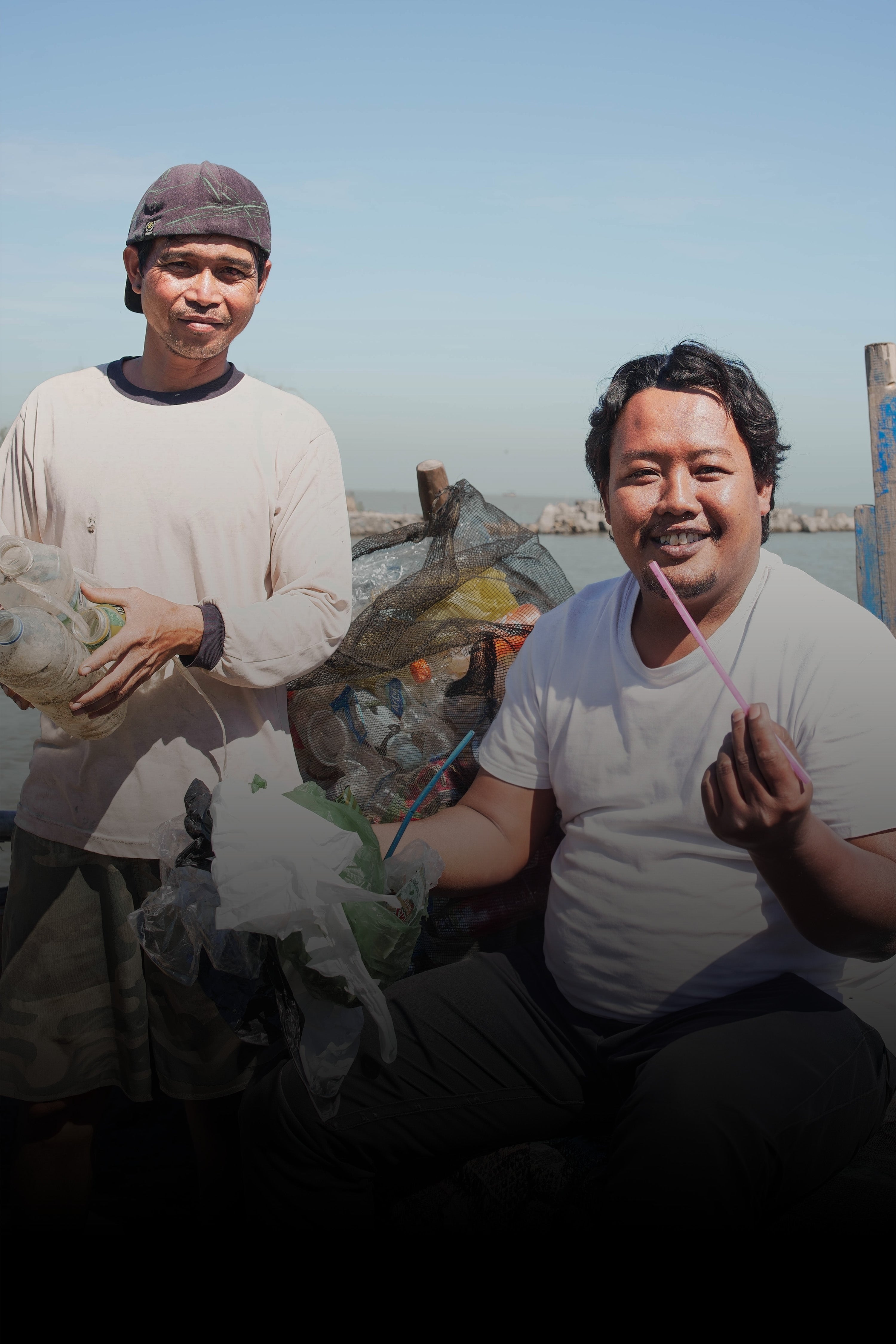 COMMUNITY LEADERS Accessoires: Zwei Männer sammeln Plastikmüll am Wasser, einer hält einen pinken Strohhalm.