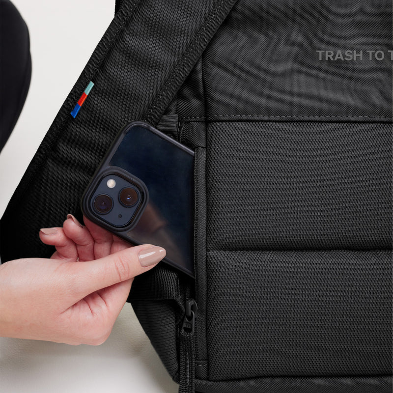 DAYPACK 2.0 Rucksack in black (Schwarz), gehalten von einer Frau, die ihr Telefon in eine Tasche steckt.