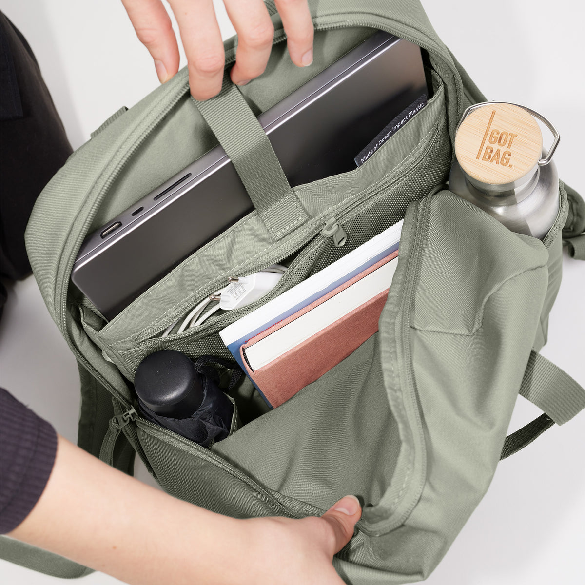 DAYPACK LOOP Rucksack in bass (Salbeigrün) wird von Händen geöffnet, zeigt Laptop, Bücher und Wasserflasche.