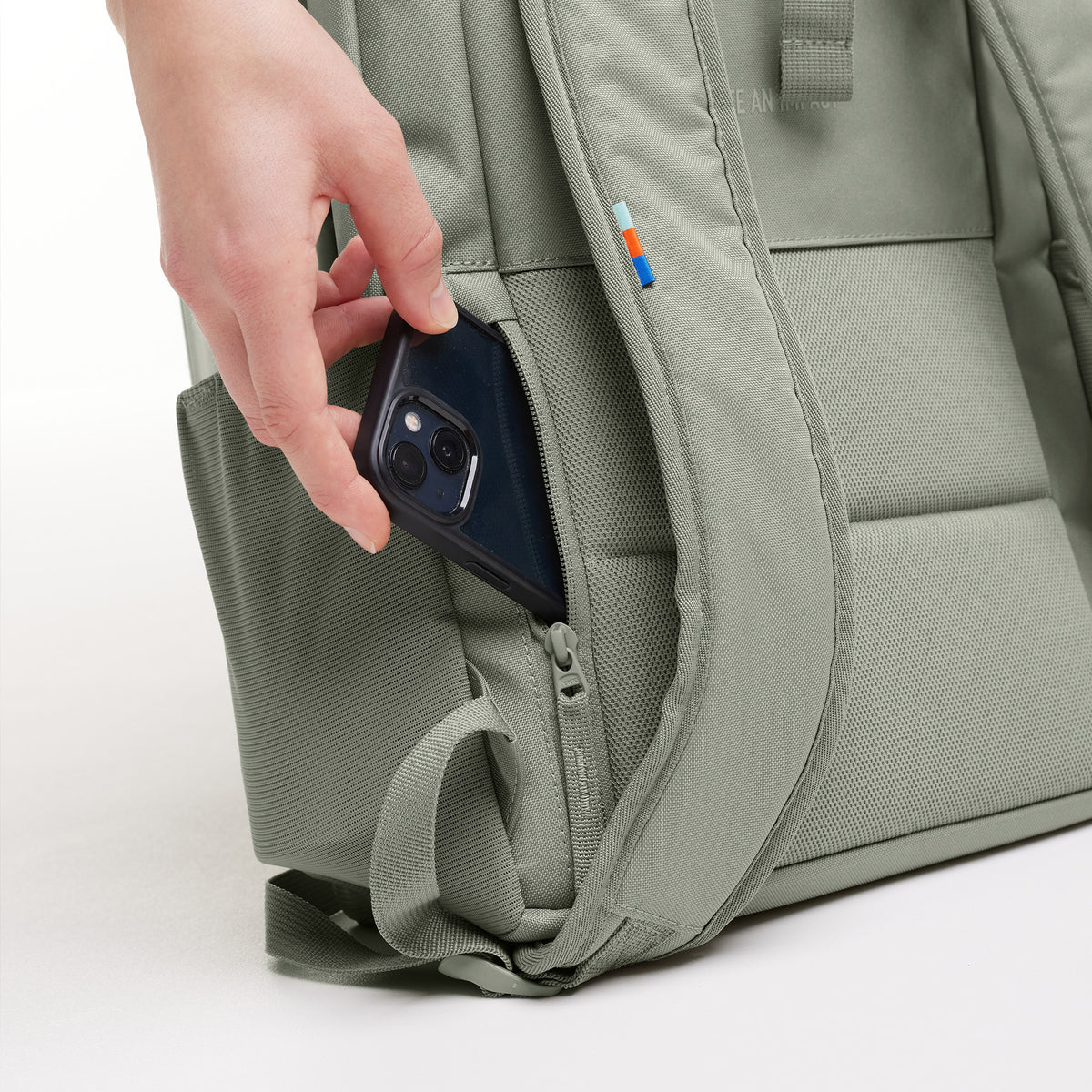 DAYPACK LOOP Rucksack in bass (Salbeigrün), eine Hand steckt ein Telefon in die Seitentasche.