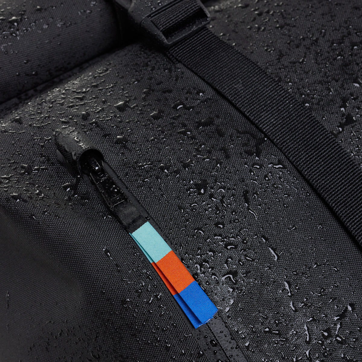 ROLLTOP LITE Rucksack, Detailaufnahme des wasserabweisenden Materials mit Regentropfen, Reißverschluss und farbigem Zugband.