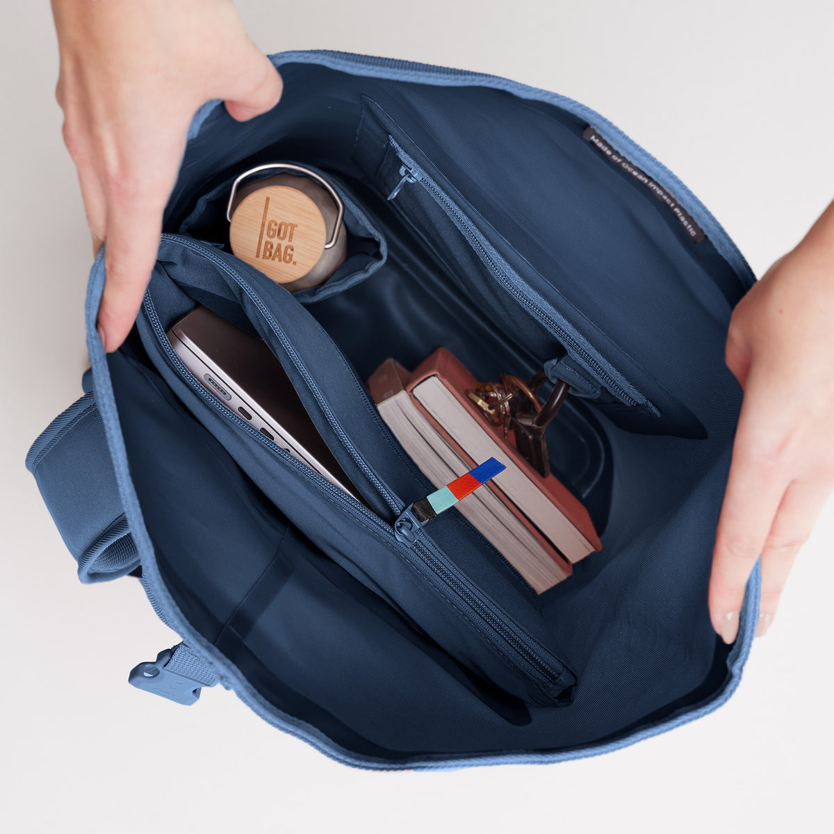 ROLLTOP LITE Rucksack in ocean, von Händen geöffnet, mit Laptop, Flasche, Büchern & Schlüssel.
