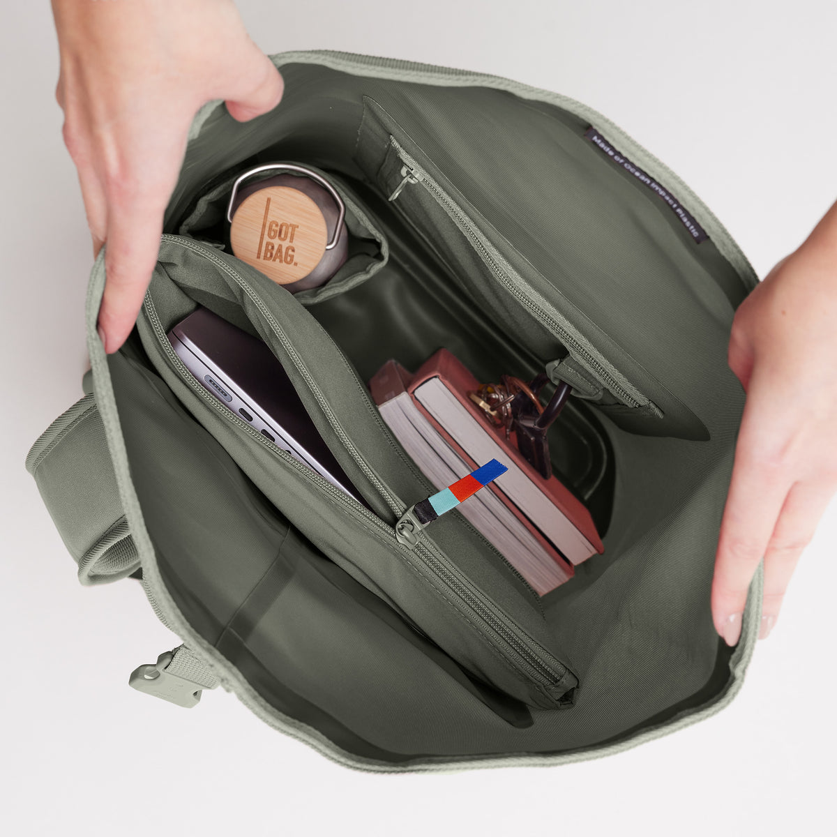 ROLLTOP Rucksack in bass (Salbeigrün) geöffnet von Händen gehalten, mit Laptop und Wasserflasche im Inneren.