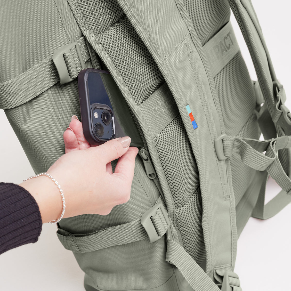 ROLLTOP 2.0 Rucksack in bass (Salbeigrün), Detailaufnahme einer Hand, die ein Smartphone in die Reißverschlusstasche steckt.