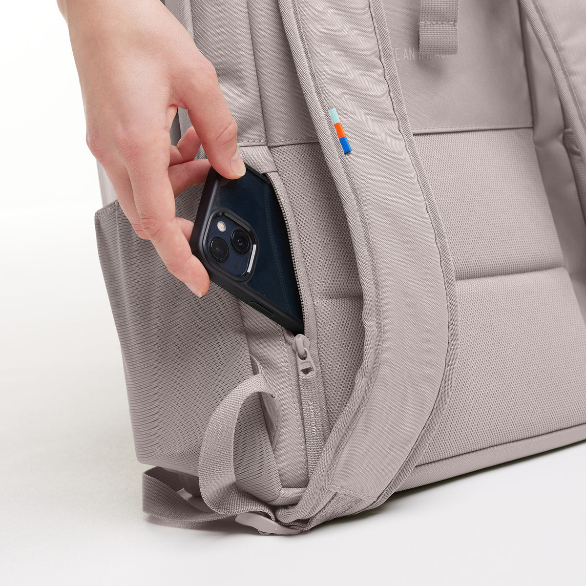 Eine Hand steckt ein Smartphone in die Seitentasche des DAYPACK LOOP Rucksacks in seahorse (graues Pastellrosa).