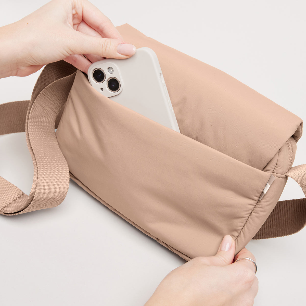 CLOUD CROSSBODY BAG Tasche in sandbar (warmes Beige mit rötlichem Unterton), ein Smartphone wird von Händen hineingesteckt.
