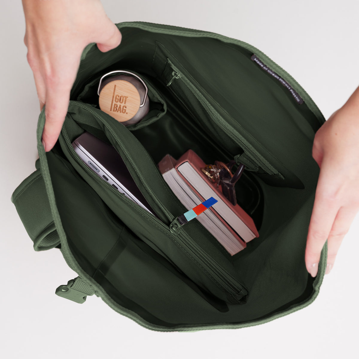 ROLLTOP LITE Rucksack in algae (Dunkelgrün mit Graustich) von Händen geöffnet, zeigt Laptop, Flasche und Bücher.
