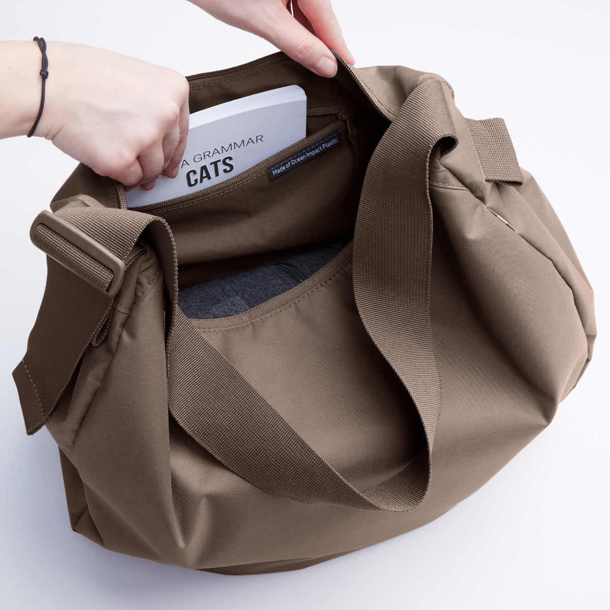 CURVED BAG Tasche in oyster (klassisches Taupe-Braun), Hand steckt Buch hinein, Made of Ocean Impact Plastic Label sichtbar.