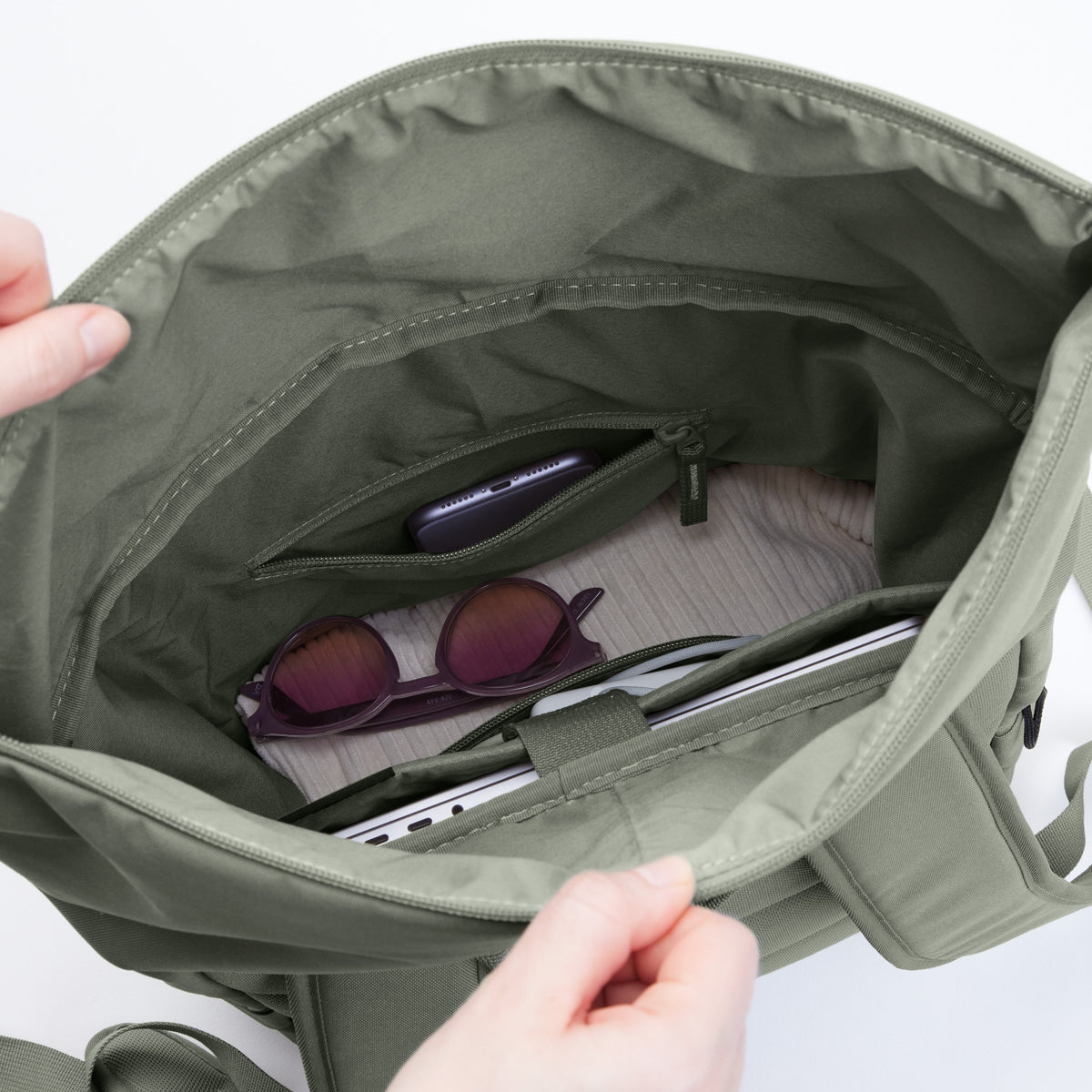 Hände öffnen den ROLLTOP EASY Rucksack in bass (Salbeigrün), Blick ins Innere mit Staufächern, Laptop, Handy.