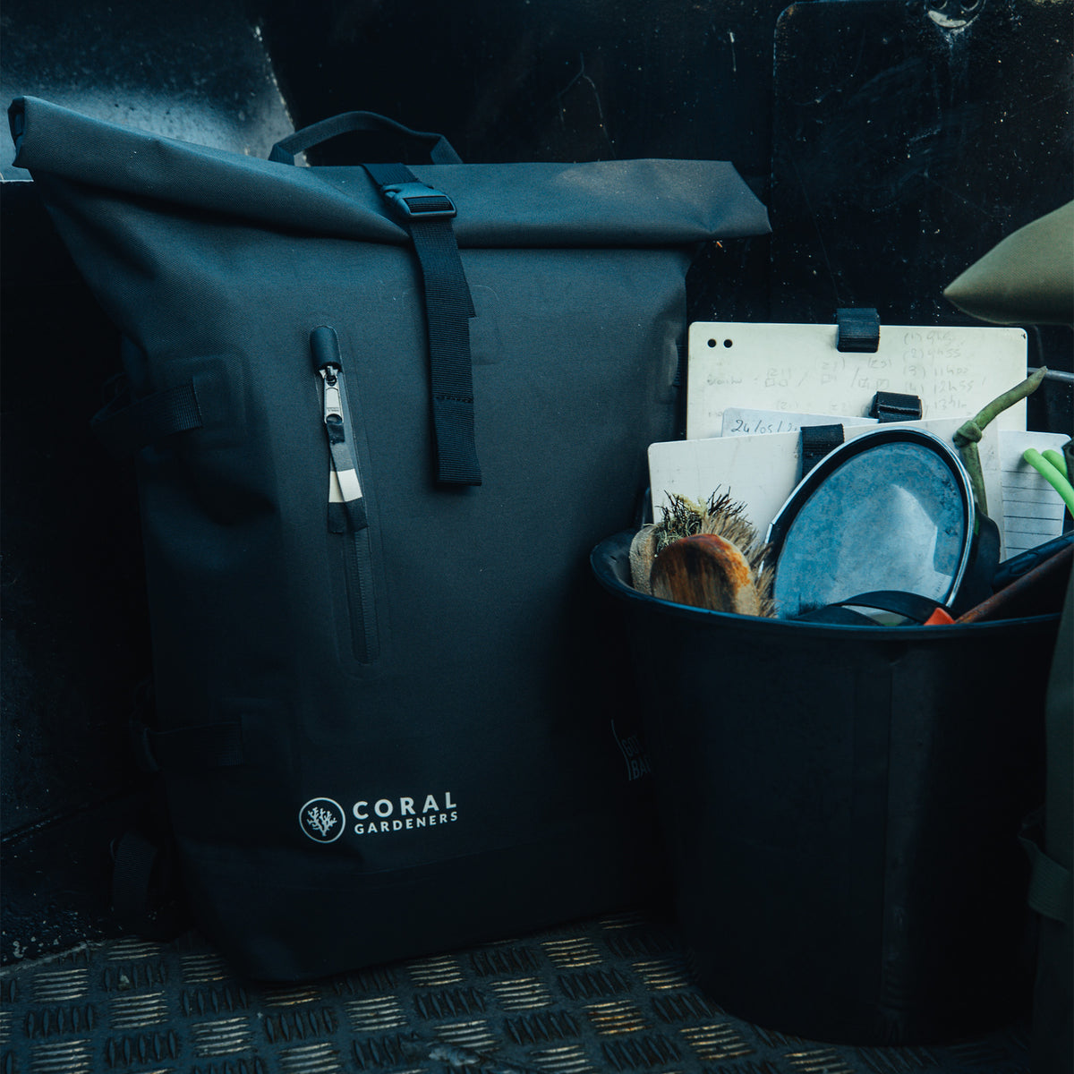 ROLLTOP Rucksack in black (Schwarz) auf dunklem Metallboden neben einem Eimer mit Tauchausrüstung.
