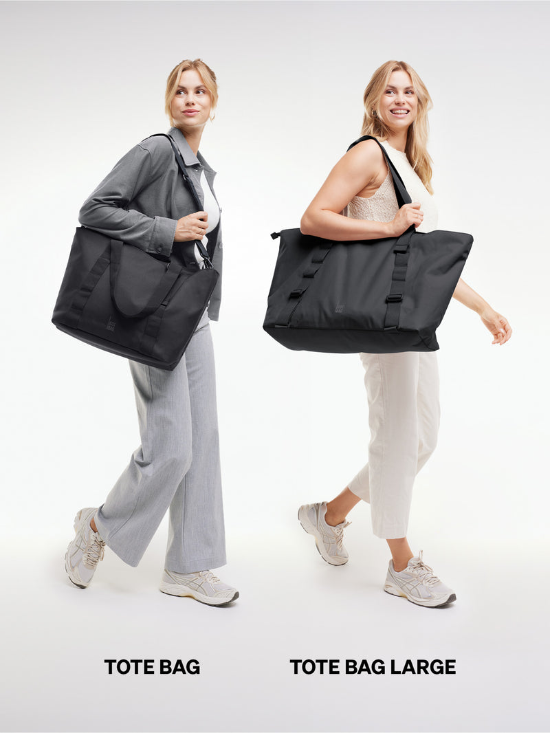 Zwei Frauen präsentieren TOTE BAG und TOTE BAG LARGE Taschen im Größenvergleich, beide über der Schulter getragen.