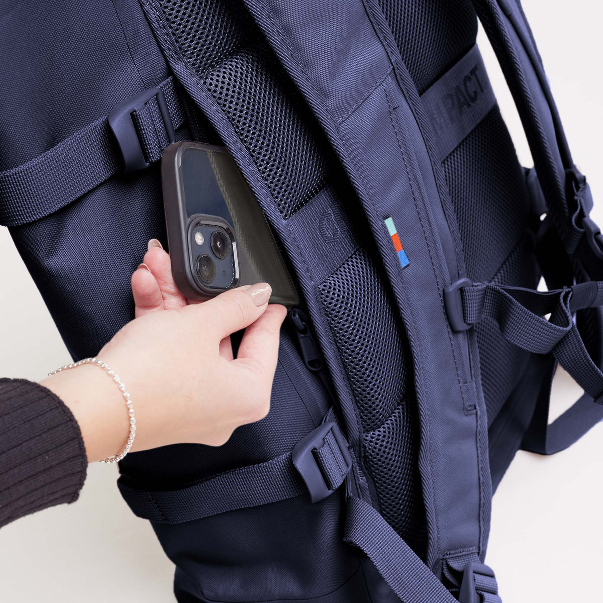 ROLLTOP 2.0 Rucksack in tide (tiefes Dunkelblau mit Lilastich), Frau steckt Smartphone in versteckte Reißverschlusstasche.