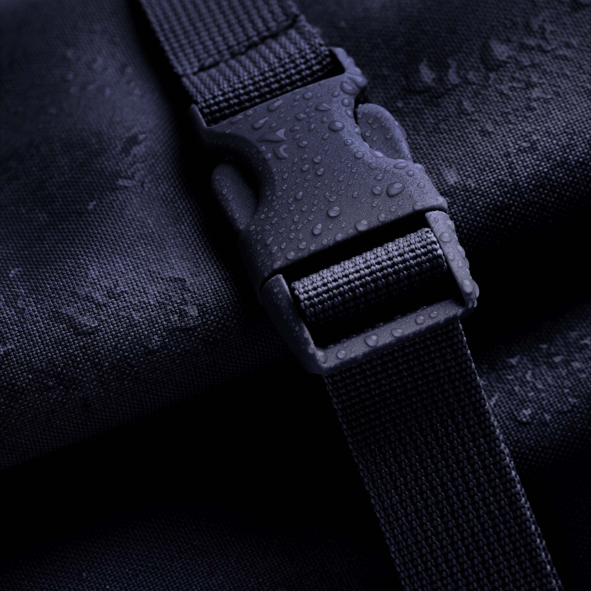 ROLLTOP Rucksack in tide (tiefes Dunkelblau mit Lilastich), Detailaufnahme der Schnalle, mit Wassertropfen bedeckt.