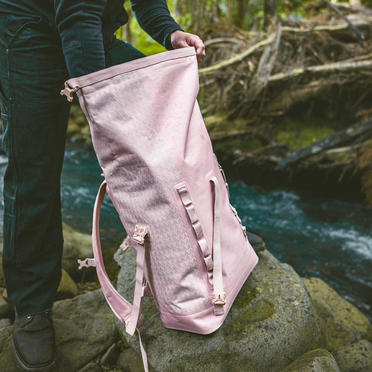 ROLLTOP ON THE ROAM Rucksack in dirty pink, von einem Mann am Flussufer auf einem moosbewachsenen Felsen angepasst.