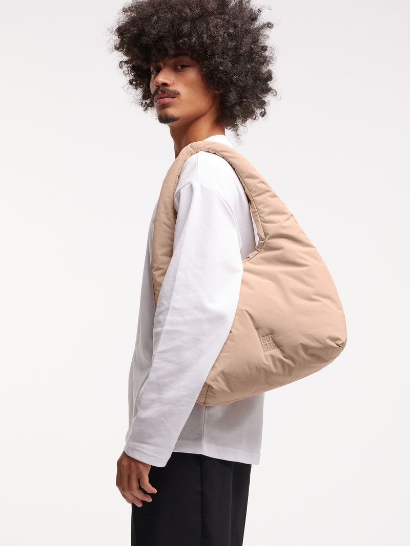CLOUD SHOULDER BAG Tasche in sandbar (warmes Beige mit rötlichem Unterton) von einem Mann über der Schulter getragen.