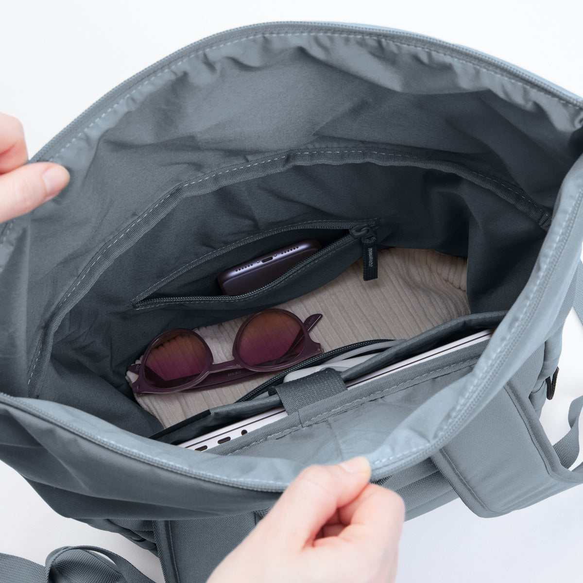 ROLLTOP EASY Rucksack in marlin (grau-blau) mit Händen geöffnet, die das Innenleben mit Handy, Sonnenbrille und Laptop zeigen.