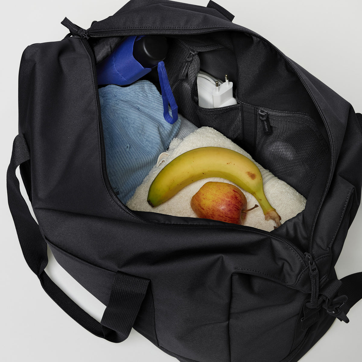 WEEKEND BAG Tasche in black (Schwarz), von oben geöffnet mit Alltagsgegenständen wie Obst und Ladegerät.