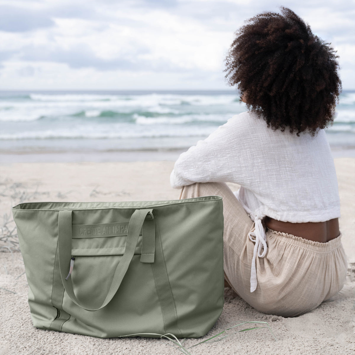 TOTE BAG Tasche in bass (Salbeigrün) liegt auf einem Sandstrand neben einer sitzenden Frau, die aufs Meer blickt.
