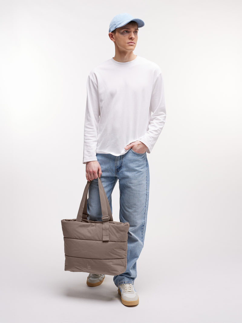 PUFFER TOTE BAG Tasche in oyster (klassisches Taupe-Braun) von einem Mann in der Hand gehalten.