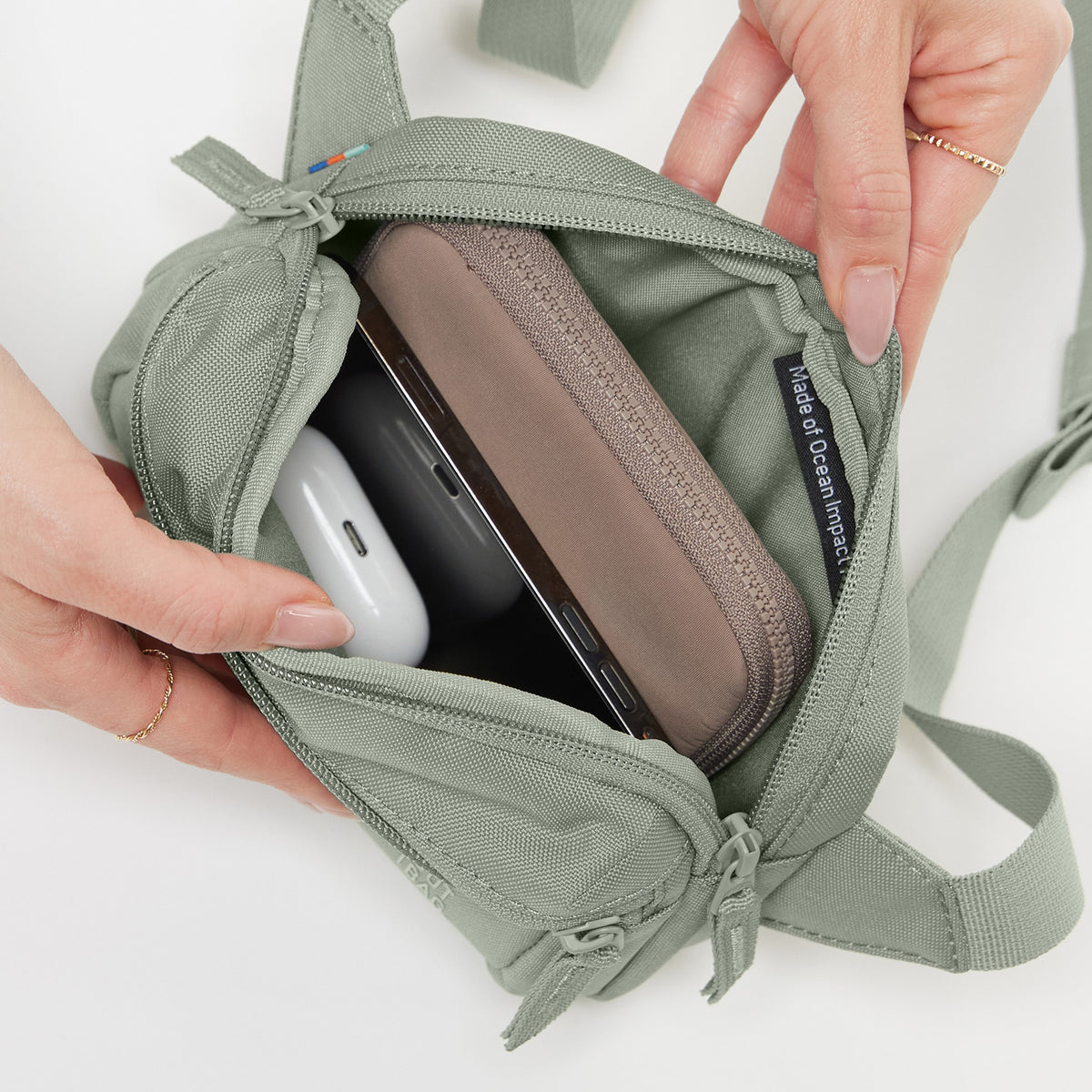 SLING BAG SMALL Tasche in bass (Salbeigrün), geöffnet von Händen, die den Inhalt zeigen.