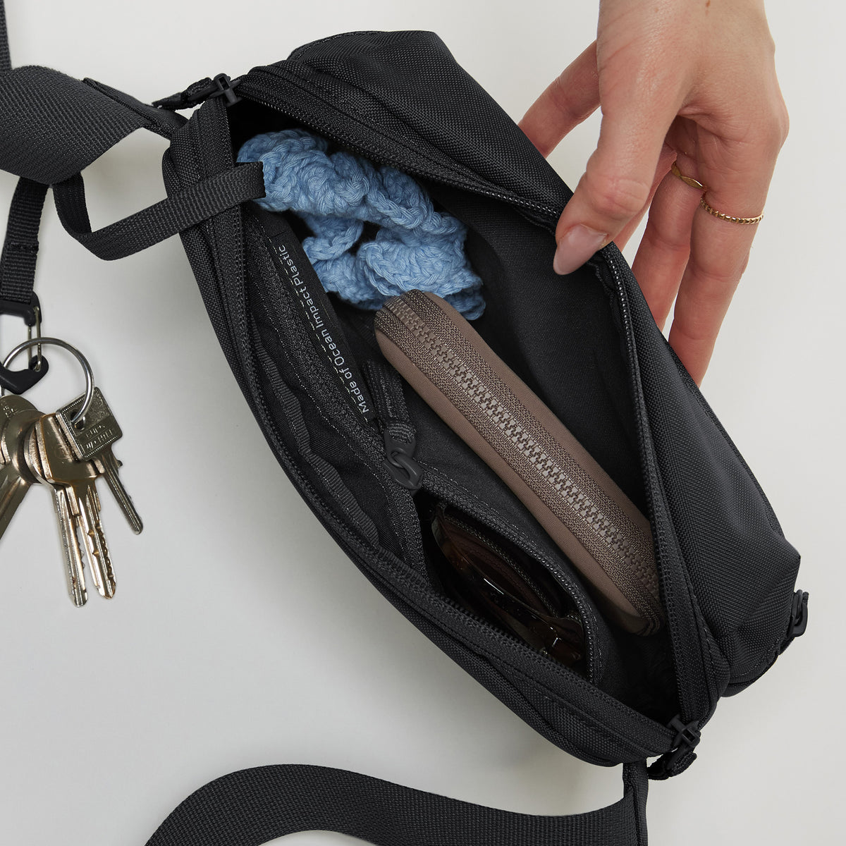 SLING BAG Tasche in black (Schwarz), geöffnet, mit einer weiblichen Hand, die hineingreift.