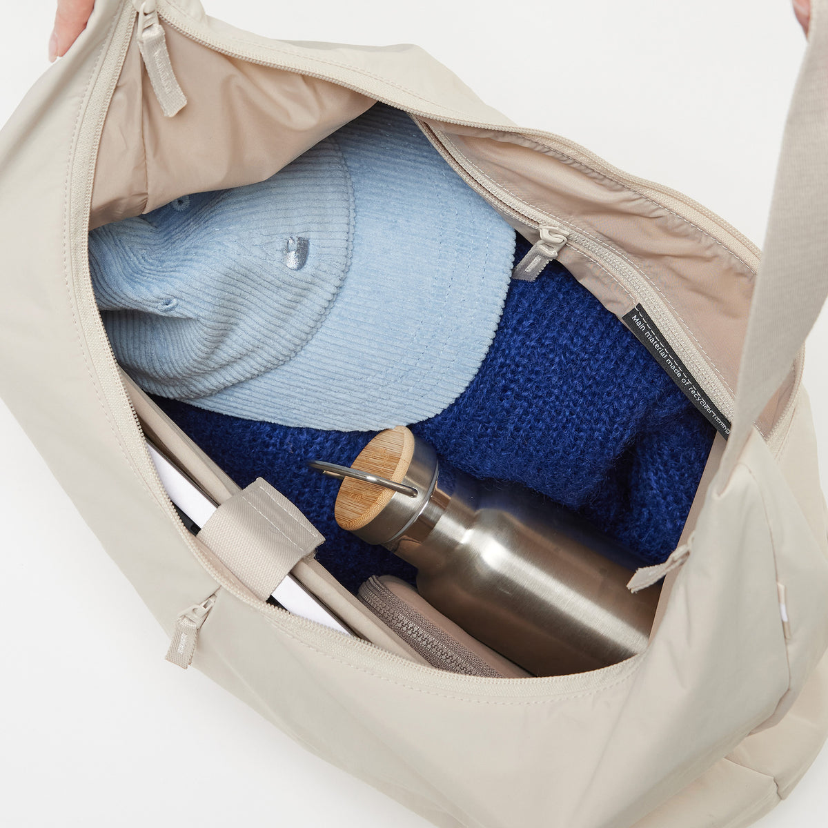 T SHAPE BAG LARGE Tasche in soft shell (warmes Cremeweiß), von oben in das Innere mit Mütze, Pullover und Trinkflasche gesehen.