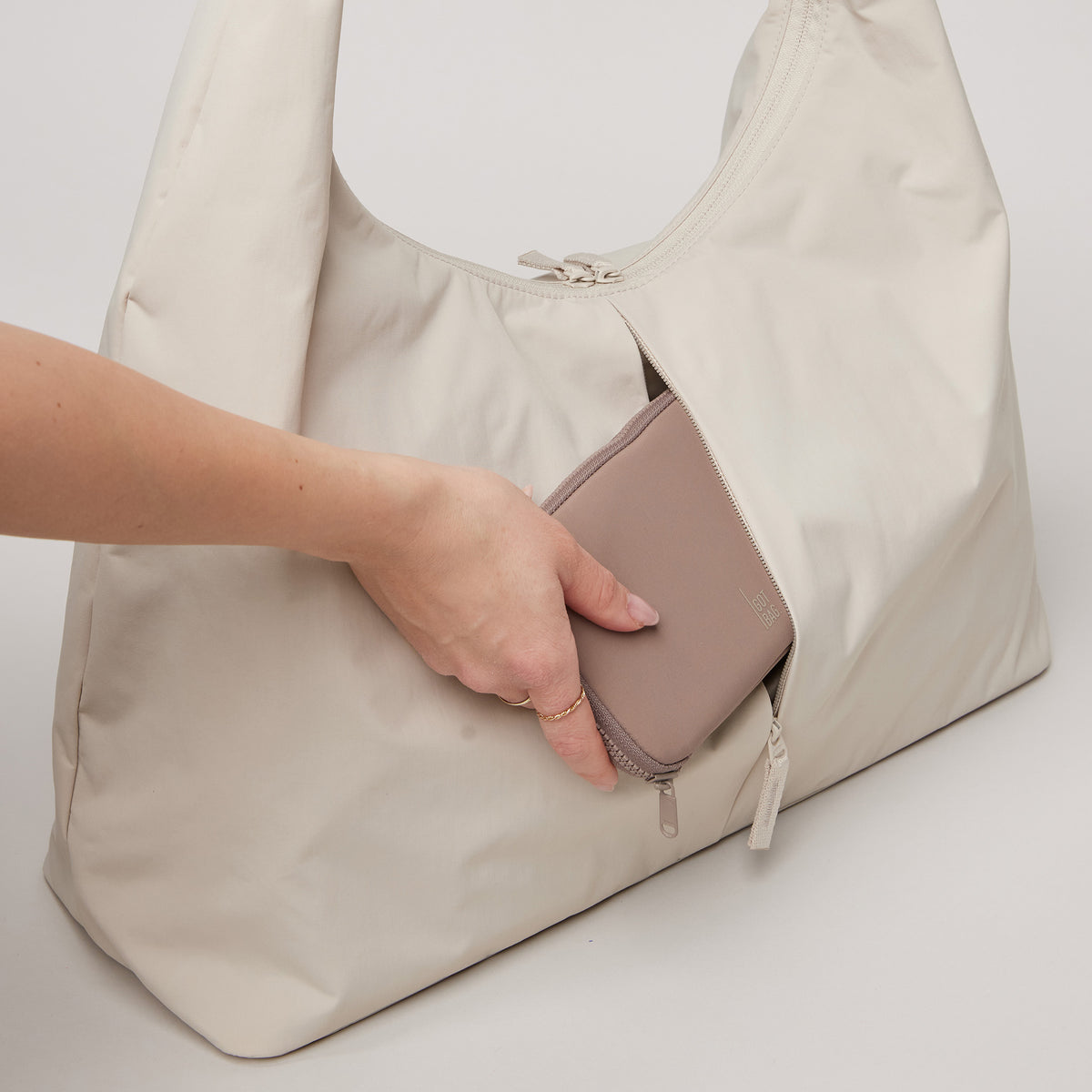 T SHAPE BAG LARGE Tasche in soft shell (warmes Cremeweiß), eine Hand steckt eine Brieftasche in die Reißverschlusstasche.