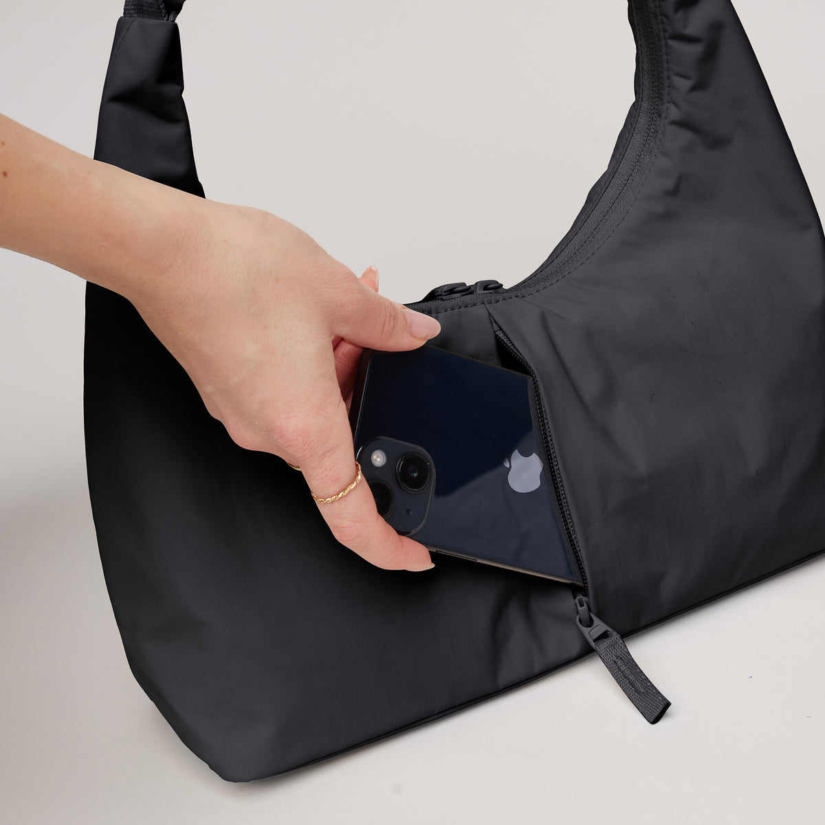 T SHAPE BAG MINI Tasche in black (Schwarz) von einer Frau, die ein Handy in die Reißverschlusstasche steckt.