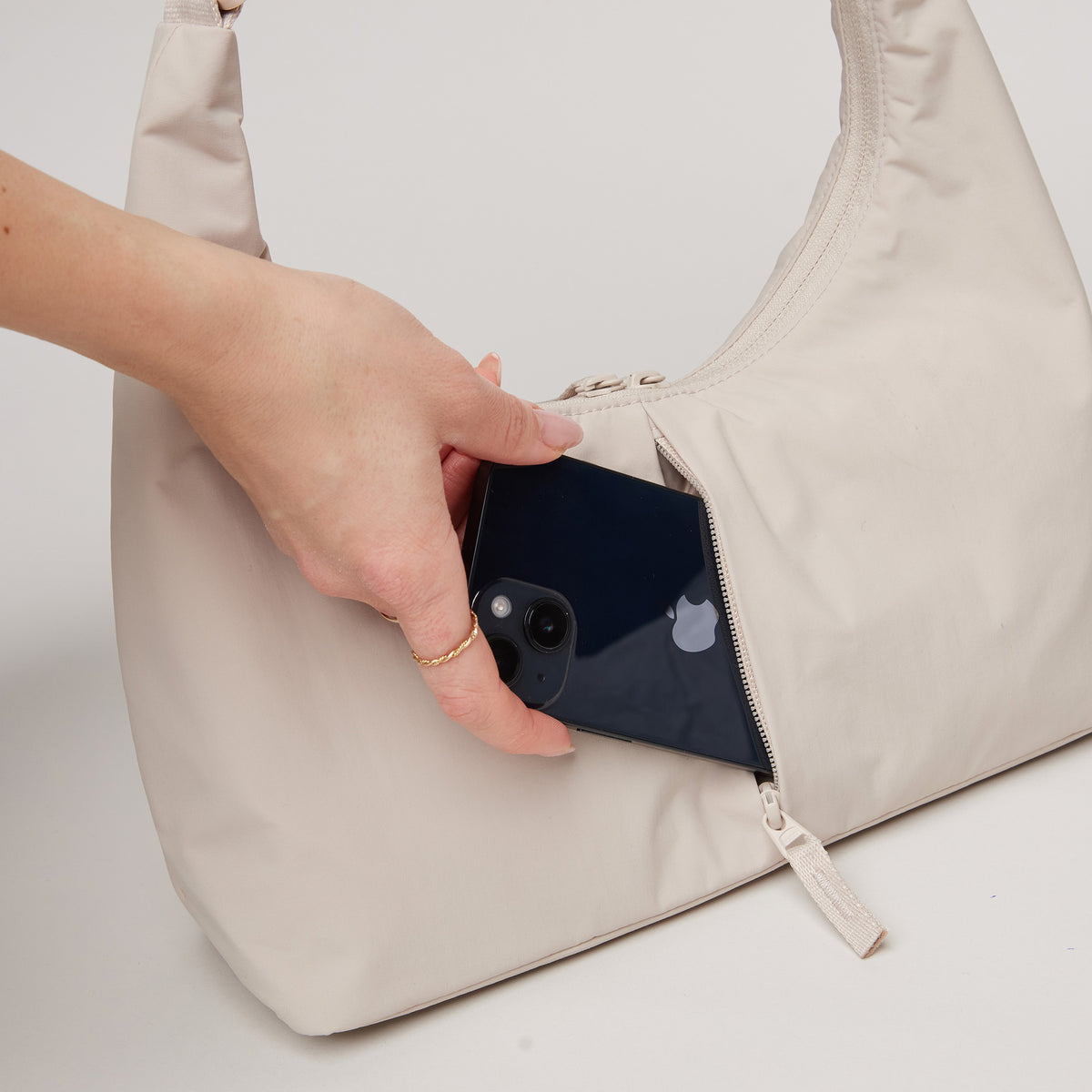 T-SHAPE BAG MINI Tasche in soft shell (warmes Cremeweiß), ein Smartphone wird von einer Frau in die Reißverschlusstasche gesteckt.