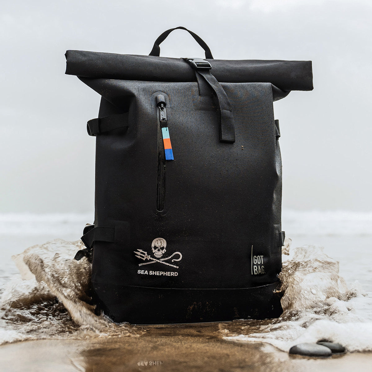 ROLLTOP Rucksack in black (Schwarz) steht am Strand im Wasser, Wellen umspülen die Basis.