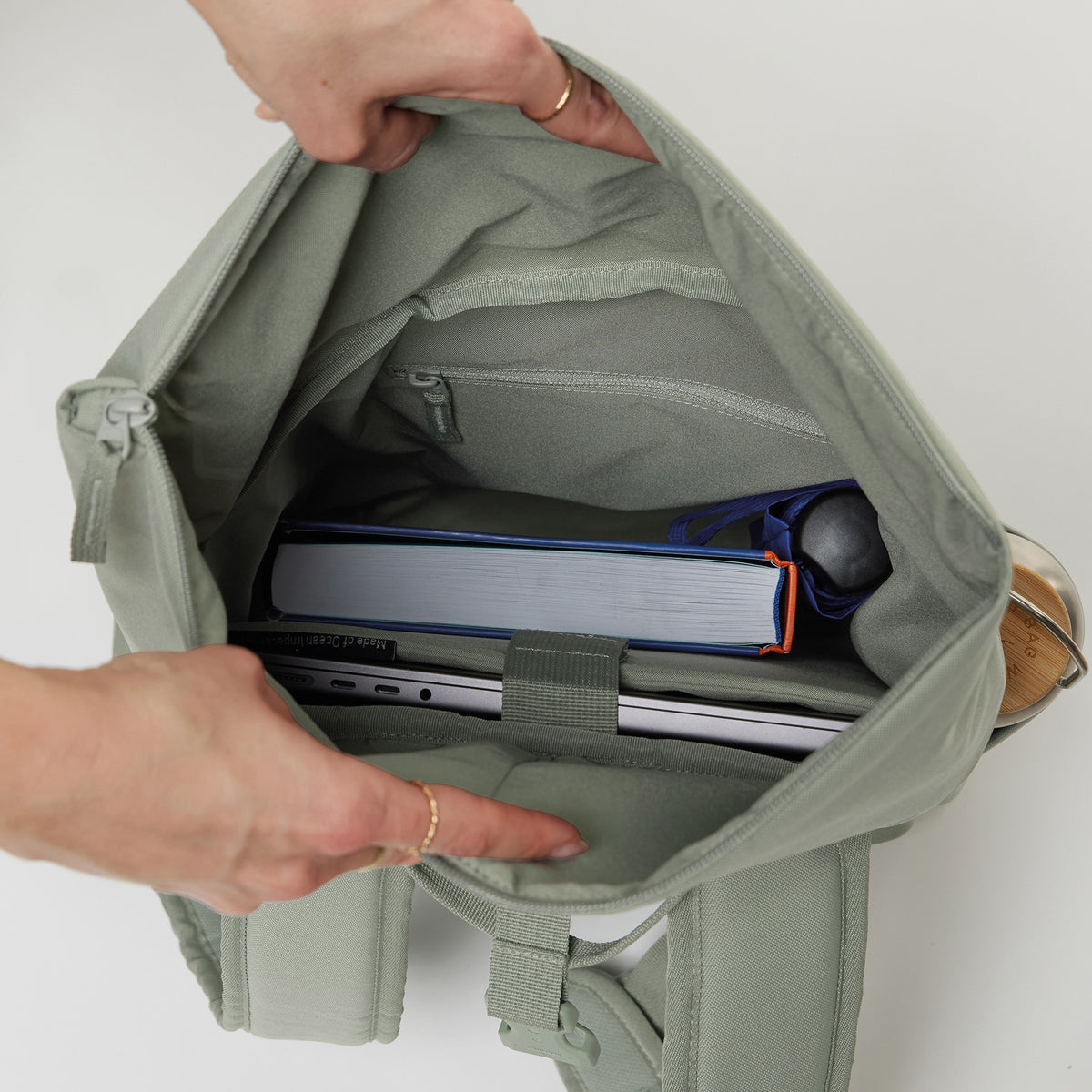 ROLLTOP EASY SMALL Rucksack in bass (Salbeigrün), Hände öffnen das Innere mit Laptop, Buch und Flasche.