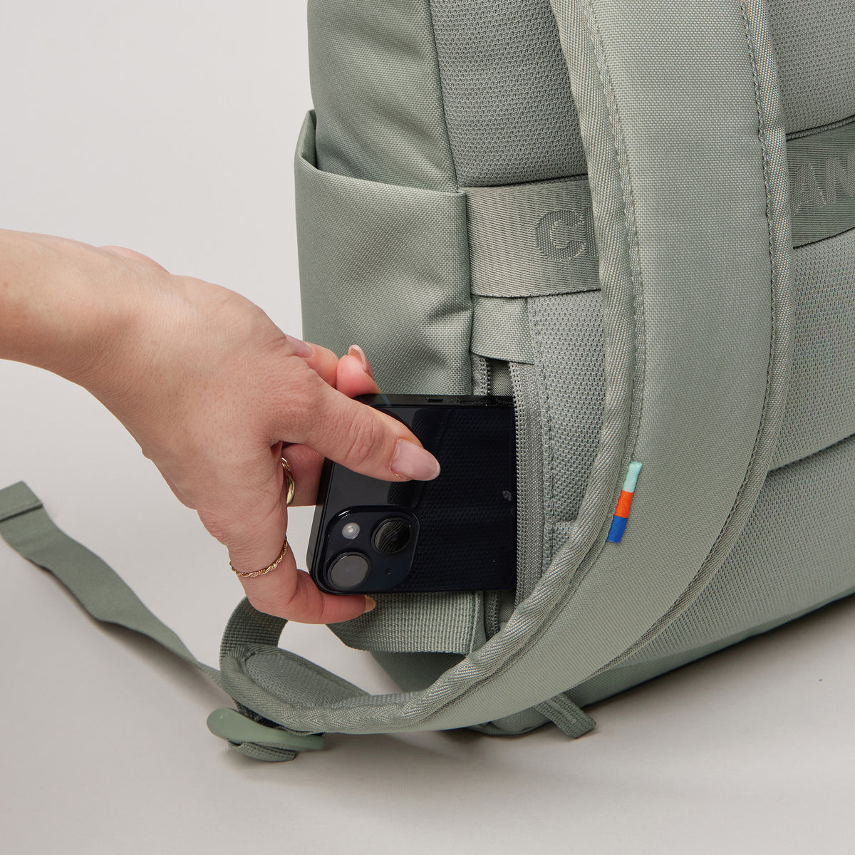 ROLLTOP EASY SMALL Rucksack in bass (Salbeigrün), mit einer Hand, die ein Smartphone in eine Seitentasche steckt.