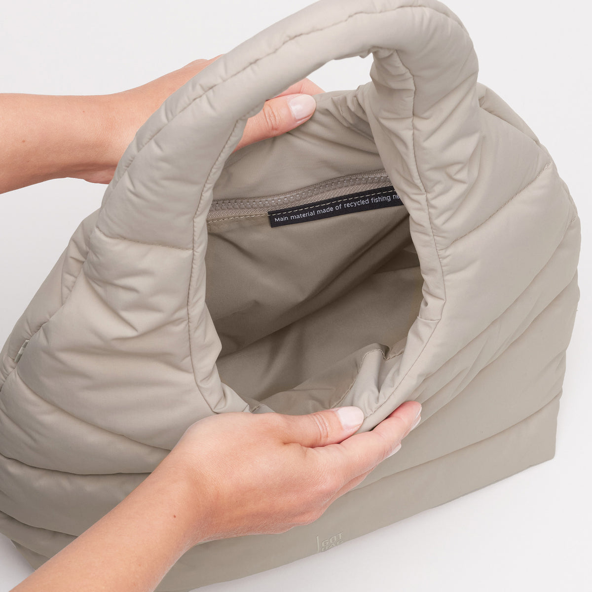 WAVY PUFFER SHOULDER BAG Tasche in scallop (helles Beige), Hände halten sie offen, mit sichtbarem Label im Inneren.