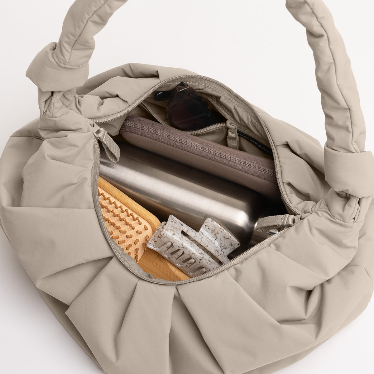 PLEAT MOON BAG LARGE Tasche in scallop (helles Beige) mit Innenansicht: Flasche, Bürste, Haarspangen und Sonnenbrille sind sichtbar.