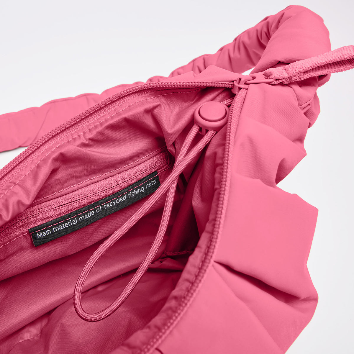 PLEAT MOON BAG SMALL Tasche in salina pink (leuchtendes Pink) als Detailaufnahme mit Aufschrift Hauptmaterial aus recycelten Fischernetzen.