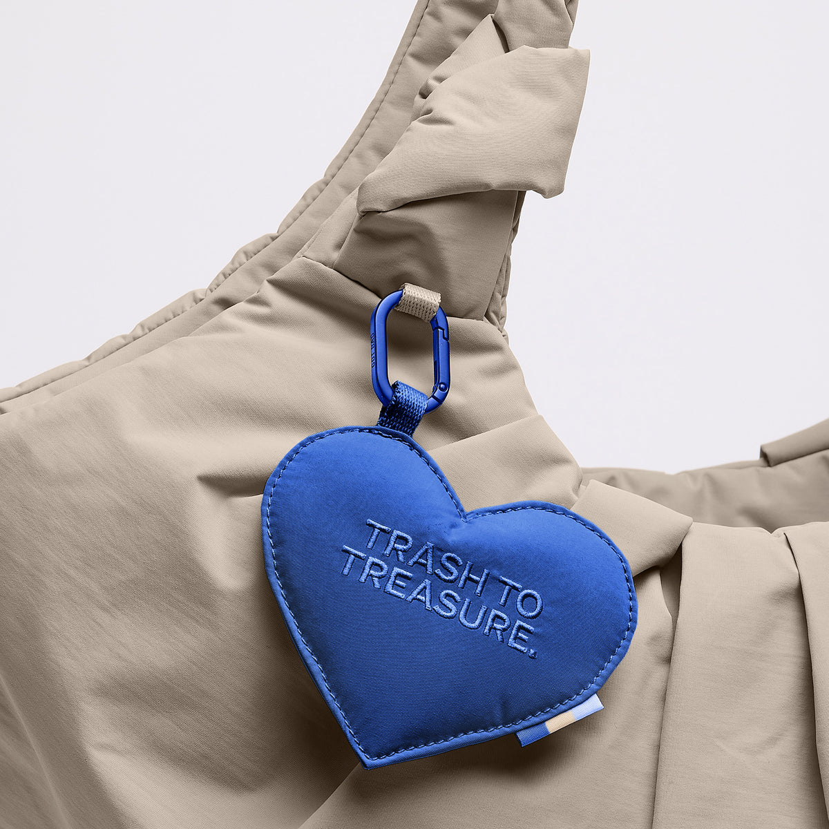 PLEAT TOTE BAG Tasche in scallop (helles Beige) mit blauem TRASH TO TREASURE Herz-Anhänger und Karabiner, Detailaufnahme.