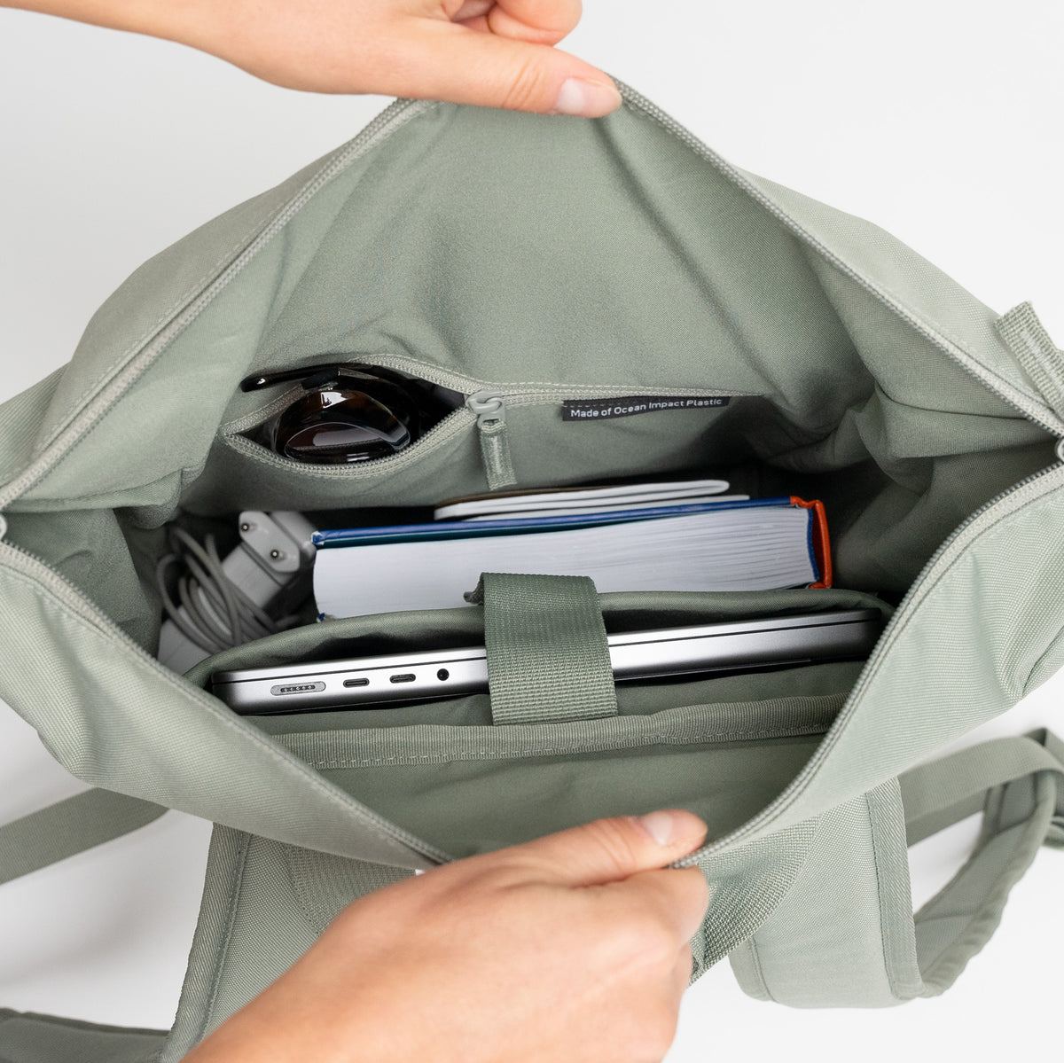 FLOW PACK Rucksack in bass (Salbeigrün) von Händen geöffnet, zeigt gut organisiertes Inneres mit Laptop und Zubehör.