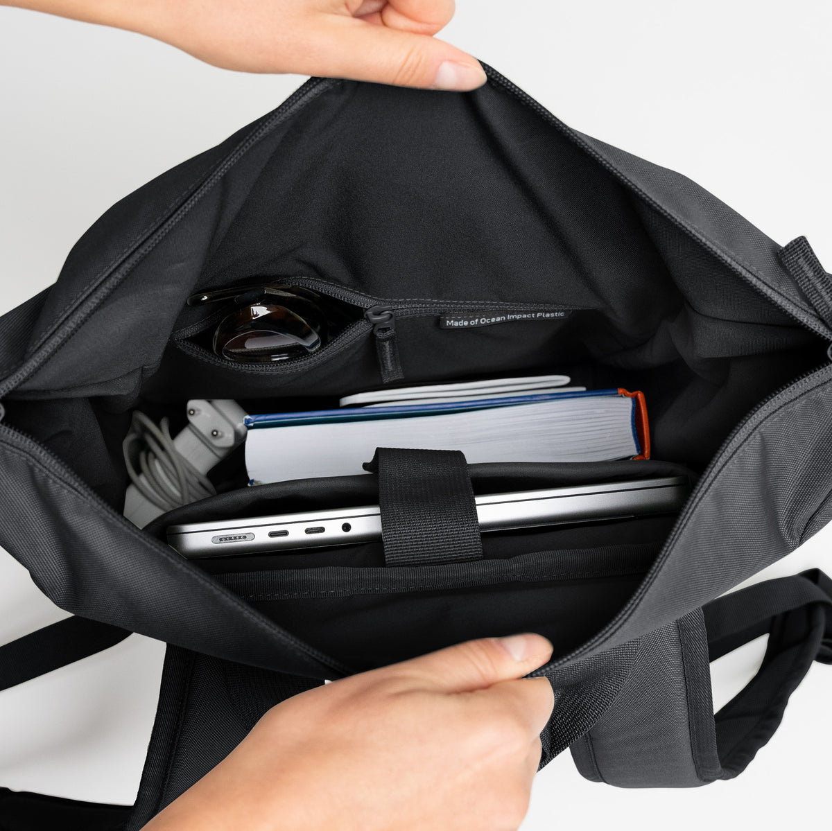 FLOW PACK Rucksack in black (Schwarz), hergestellt aus Ocean Impact Plastic, von Händen geöffnet, zeigt organisierten Innenraum.