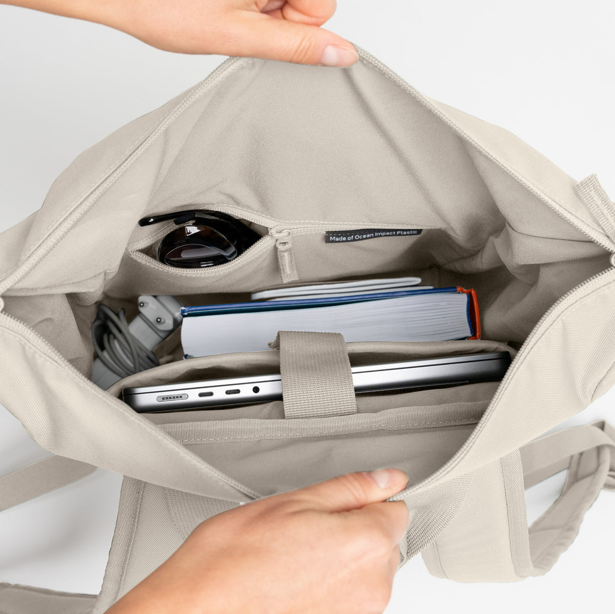 Hände öffnen FLOW PACK Rucksack in soft shell (warmes Cremeweiß) mit Blick auf den Inhalt: Laptop, Buch, Kabel.
