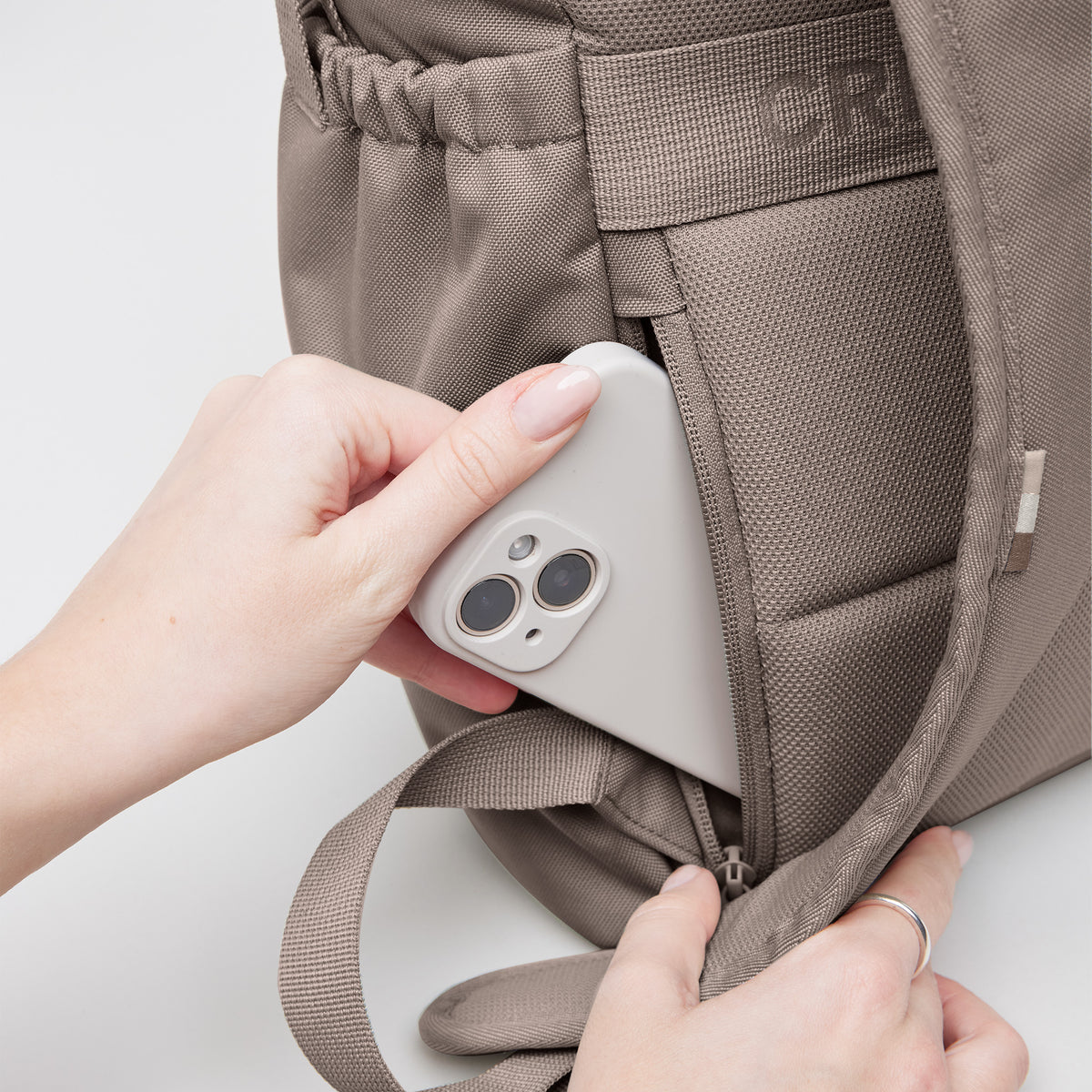 FLAP PACK MONOCHROME EDITION Rucksack in oyster (klassisches Taupe-Braun), Detailaufnahme, eine Frau steckt Handy in Geheimfach.