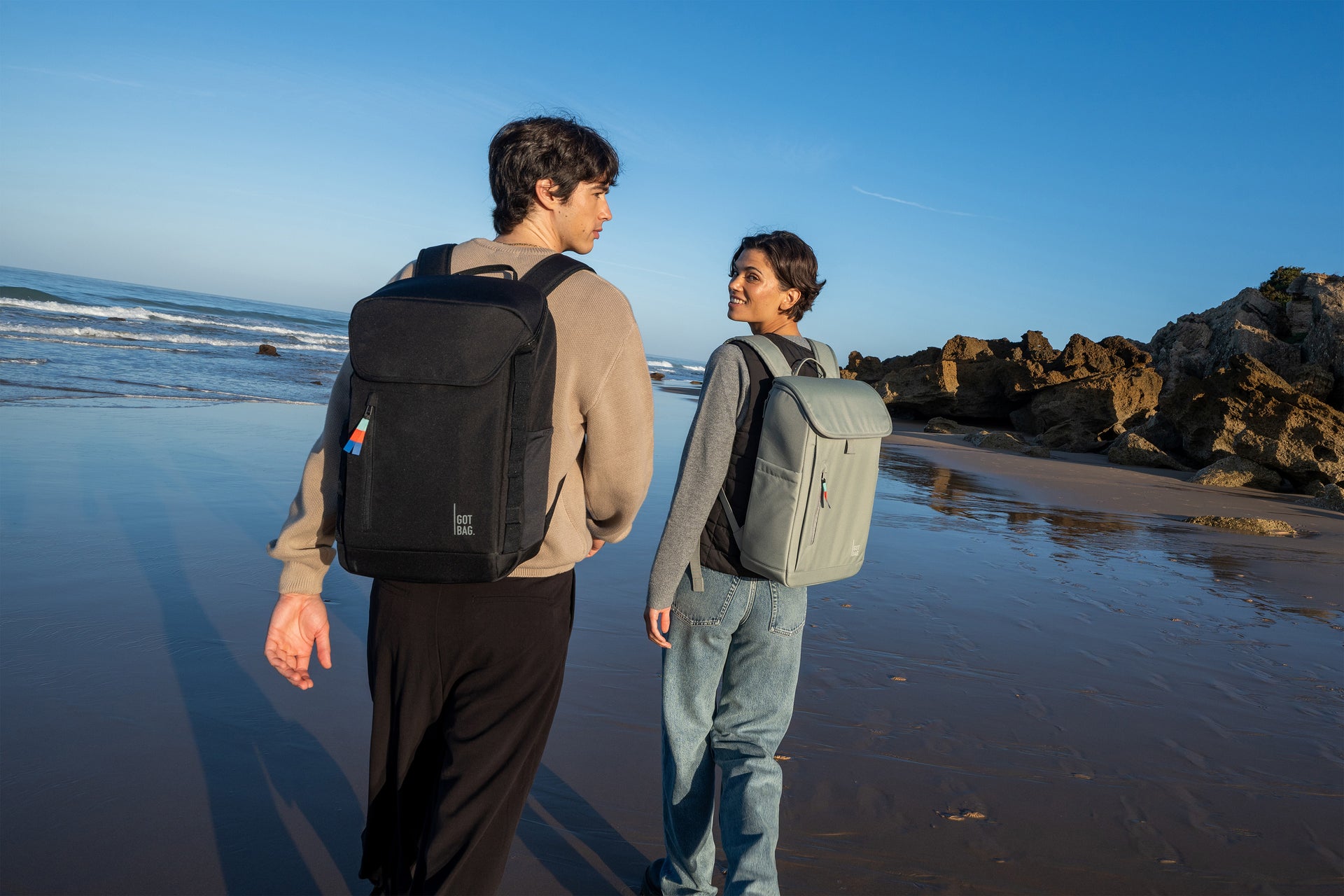 Two backpacks: PRO PACK in black (classic black) and SERENE PACK in bass (sage green), worn by man and woman.