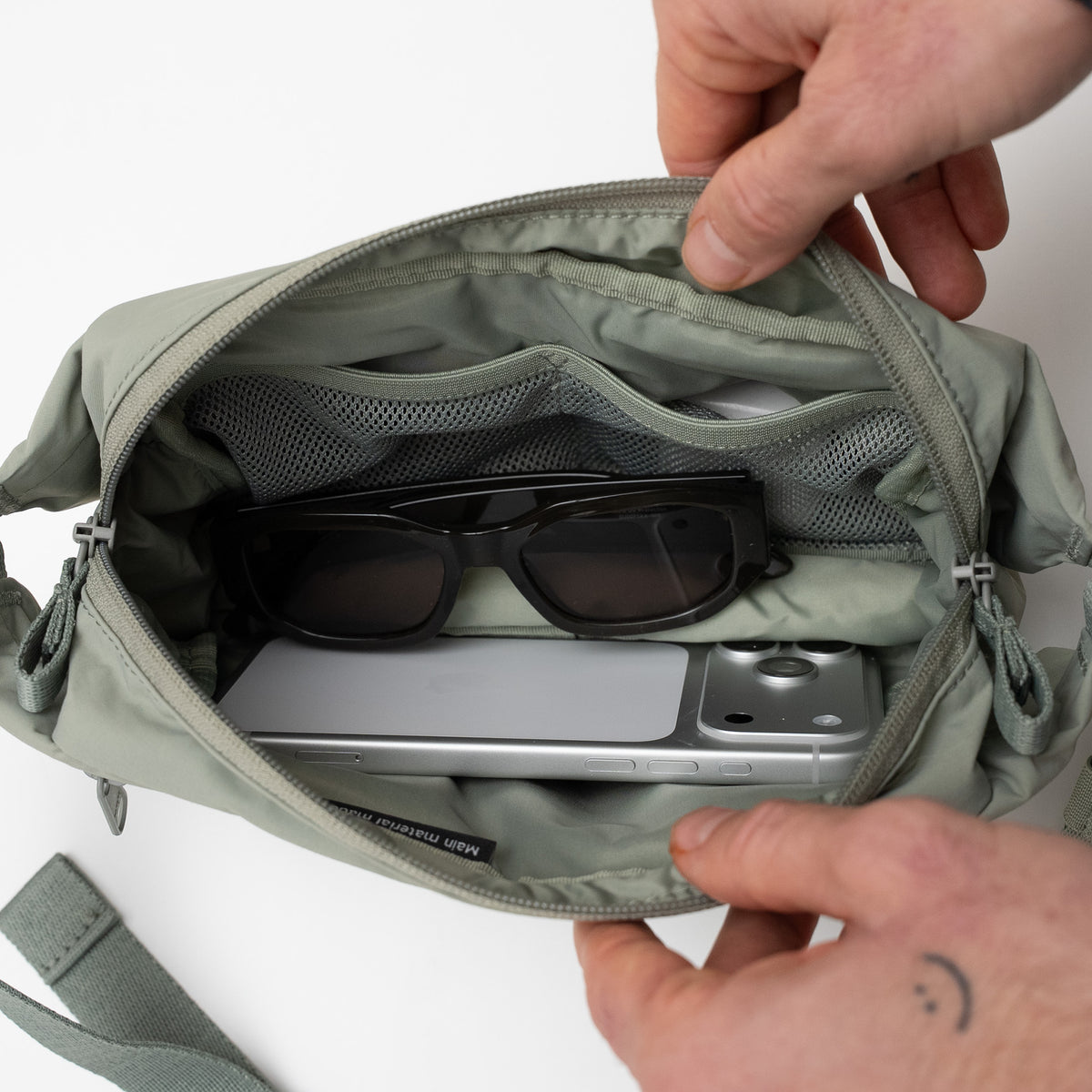 ADAPT SLING BAG Tasche in bass (Salbeigrün), geöffnet von Händen, mit Smartphone und Sonnenbrille innen.