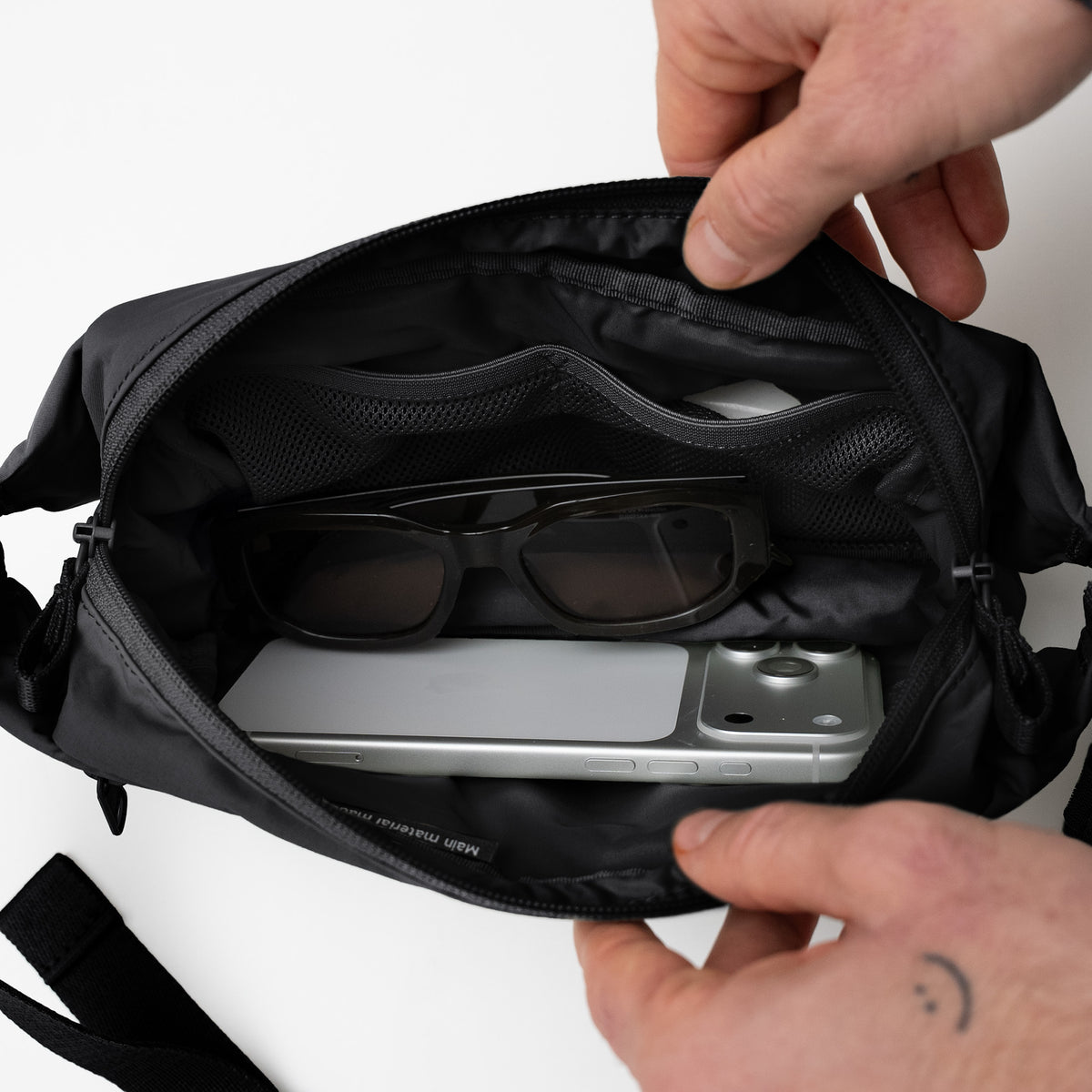 ADAPT SLING BAG Tasche in black (Schwarz) von einem Mann geöffnet zeigt organisiertes Inneres mit Brille und Smartphone.