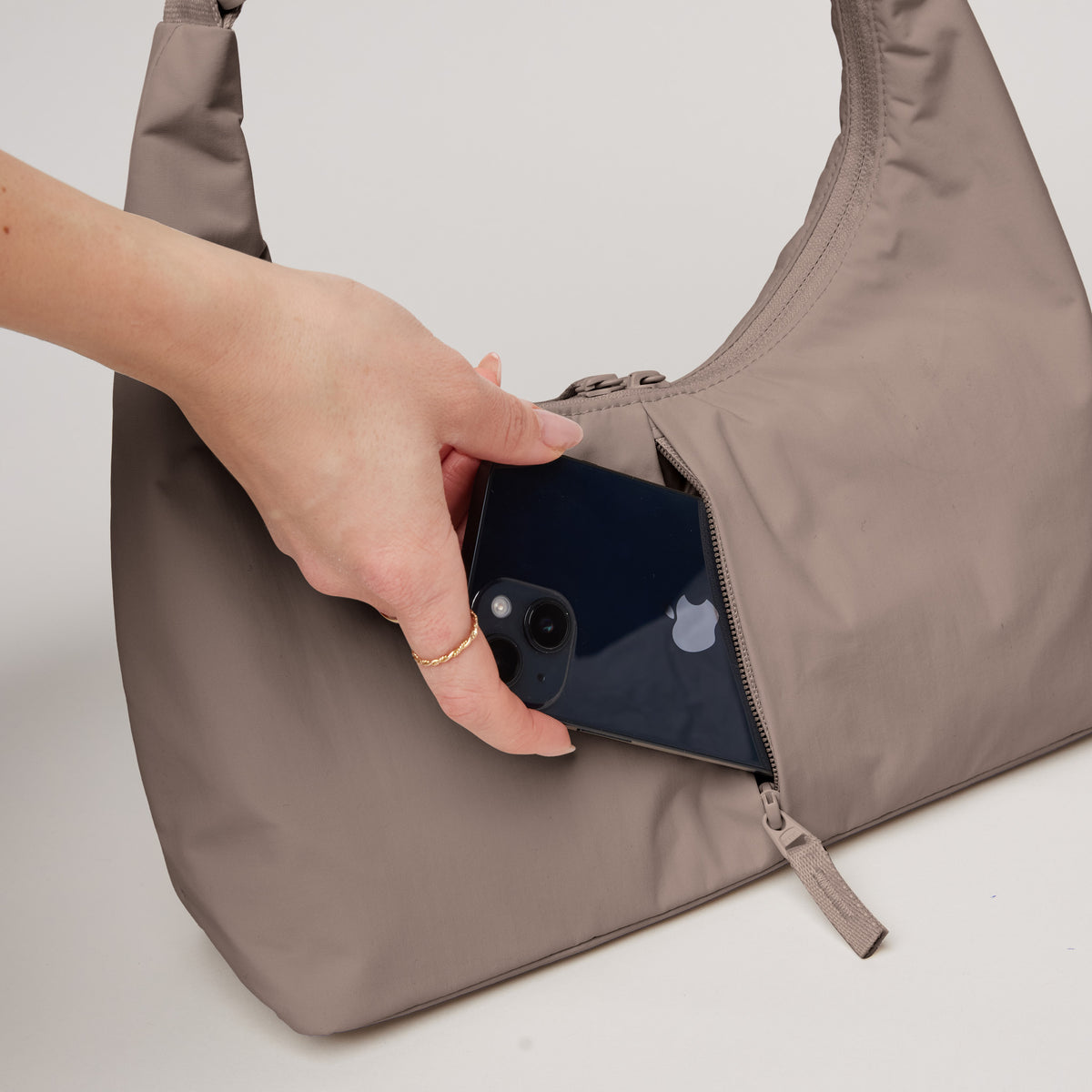 T-SHAPE BAG MINI Tasche in oyster (klassisches Taupe-Braun), eine Hand steckt Smartphone in Reißverschluss-Geheimfach.