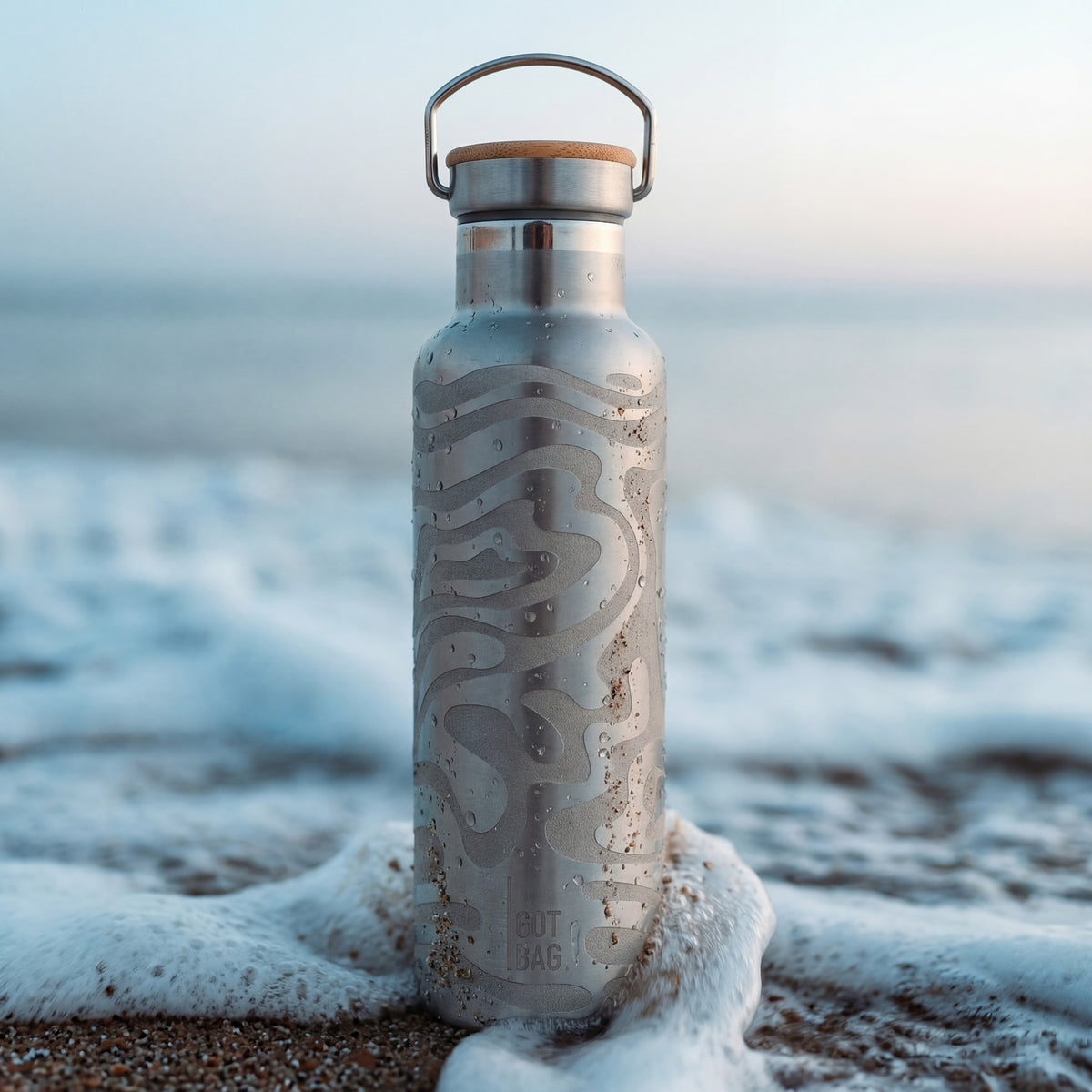 BOTTLE Accessoire, von Meereswellen umspült am Sandstrand, mit Wassertropfen und Sandkörnern.