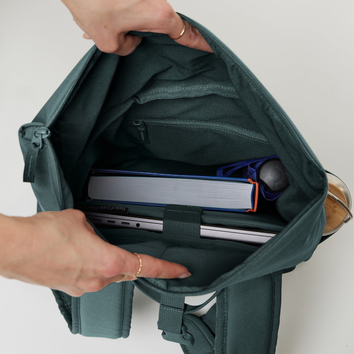 Hände öffnen ROLLTOP EASY SMALL Rucksack in sea teal (kühles Türkisgrün), gefüllt mit Laptop und Buch.