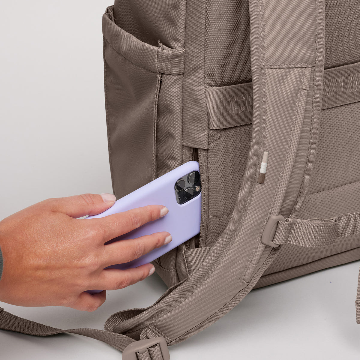 SERENE PACK Rucksack in oyster (klassisches Taupe-Braun) mit einer Frau, die ein Handy in die geheime Seitentasche steckt.